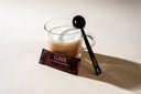 A frothy GAYA Functional Mushroom Blend cacao drink in a glass mug, with a single-serve sachet and branded measuring spoon on a beige surface