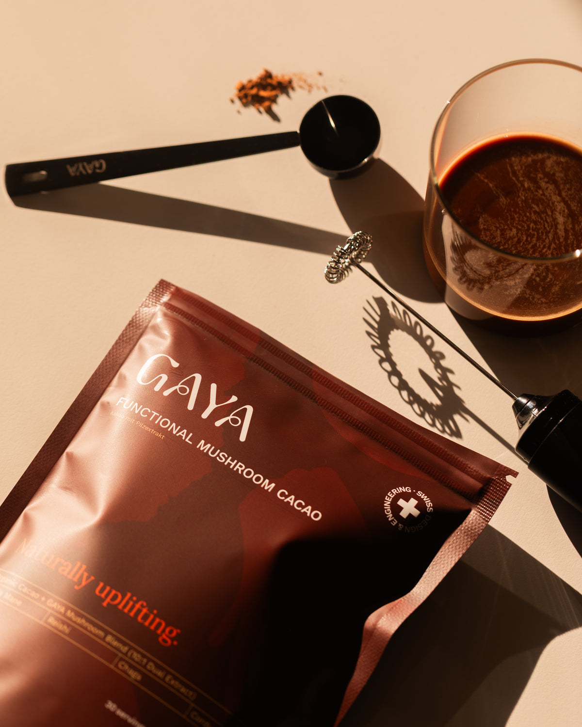 Gaya Functional Mushroom Cacao pouch with a glass of dark liquid, measuring scoop and frother, on a beige surface, demonstrating easy preparation of functional mushroom cacao